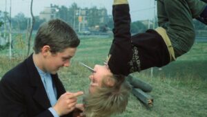 Two young men outdoors; one sits facing the other, who is hanging upside down by a harness, while the first points and holds a thin object near the other’s mouth.