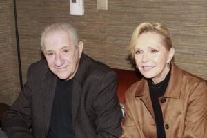 Smiling elderly man and woman sit together on a bench in a warmly striped room background.