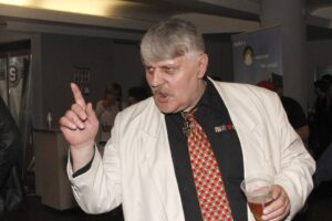 Man in a white blazer and colorful tie points finger while holding a beer at a lively event.