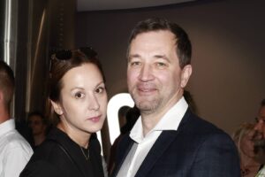 A man in a dark blazer and white shirt stands beside a woman in a black top at an indoor event, posing for a photo.