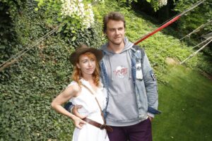 Two people posing outdoors in a garden: woman in a white dress and hat, man in a denim jacket and gray hoodie smile at the camera.