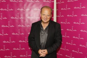 Man in a black suit posing in front of a pink backdrop with repeating white logos that read 'Dívka roku' and silhouette designs.