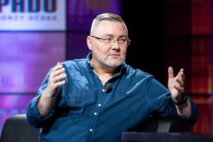 Man with gray beard and glasses speaking on a stage, wearing a blue shirt and a lapel mic, gesturing with hands.