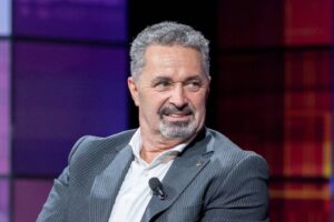 Smiling middle-aged man in a pinstripe suit on stage, microphone clipped to his lapel.