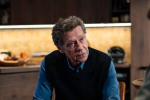 A mature man with gray hair sits at a kitchen table, wearing a blue shirt and black sweater vest, looking pensively to his left.