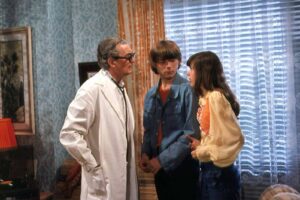 Doctor in a white coat and glasses speaks with a teenage couple in a retro living room setting.