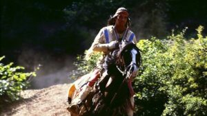 Rider in fringed Native-inspired attire on a galloping horse along a dirt trail, greenery in the background