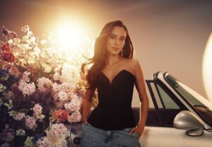 Woman in a black strapless top and jeans leaning on a convertible beside a colorful flower display with warm sunset light behind her.