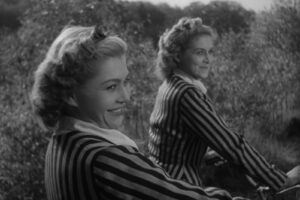 Two women in striped jackets ride bicycles in a grassy field, smiling at the camera in a black‑and‑white photo.