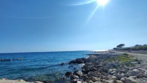 Sunny coastal scene with a rocky shore, calm blue sea, and bright sun over a clear horizon; people swim and float near the left.