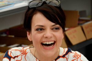Woman with dark hair laughing, sunglasses resting on top of her head in an indoor setting.