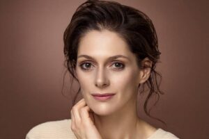Portrait of a woman with brown hair pulled back, touching her chin gently against a warm brown backdrop.