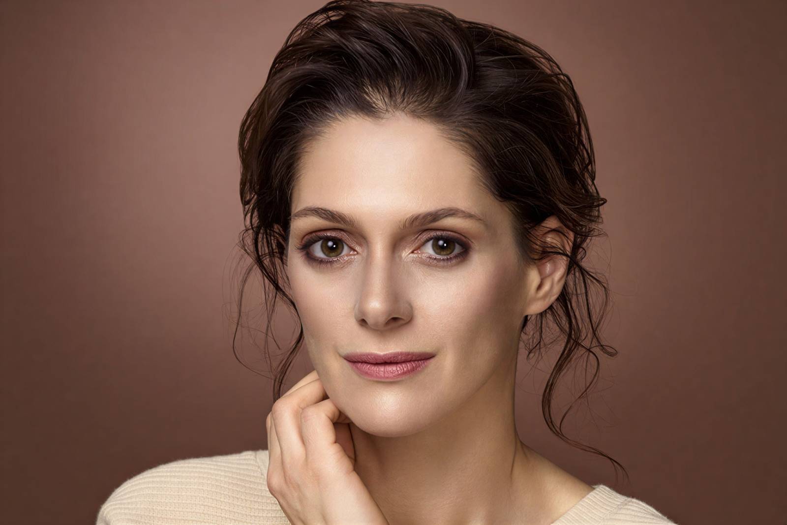 Portrait of a woman with brown hair pulled back, touching her chin gently against a warm brown backdrop.