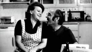 A smiling couple sits close together in a kitchen, sharing a warm moment as they look at each other.