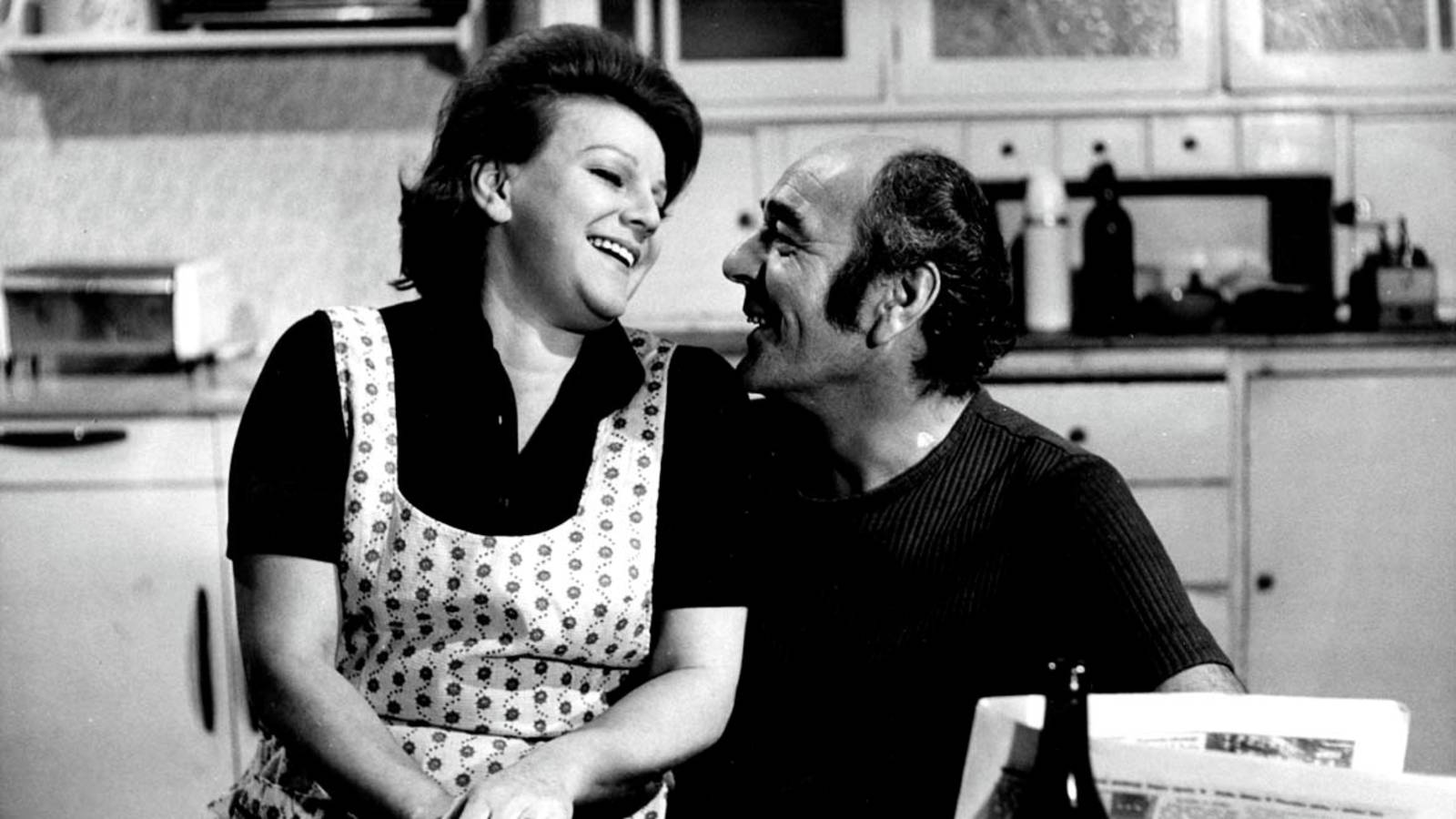 A smiling couple sits close together in a kitchen, sharing a warm moment as they look at each other.