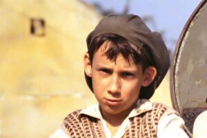 Close-up of a young boy wearing a brown cap and patterned vest, looking toward the camera outdoors in a sunny setting.