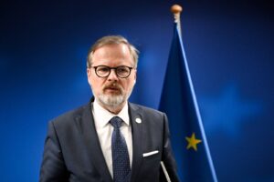 Man in a dark suit and round glasses delivering a speech, with a European flag in the blue background.