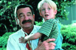 Older man with a mustache holding a young blond boy in a striped shirt outdoors in a garden setting.