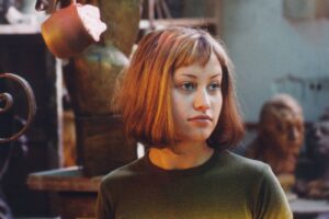 Young woman with a shoulder-length brown bob, olive-green top, looking to the right; blurred sculpture background.