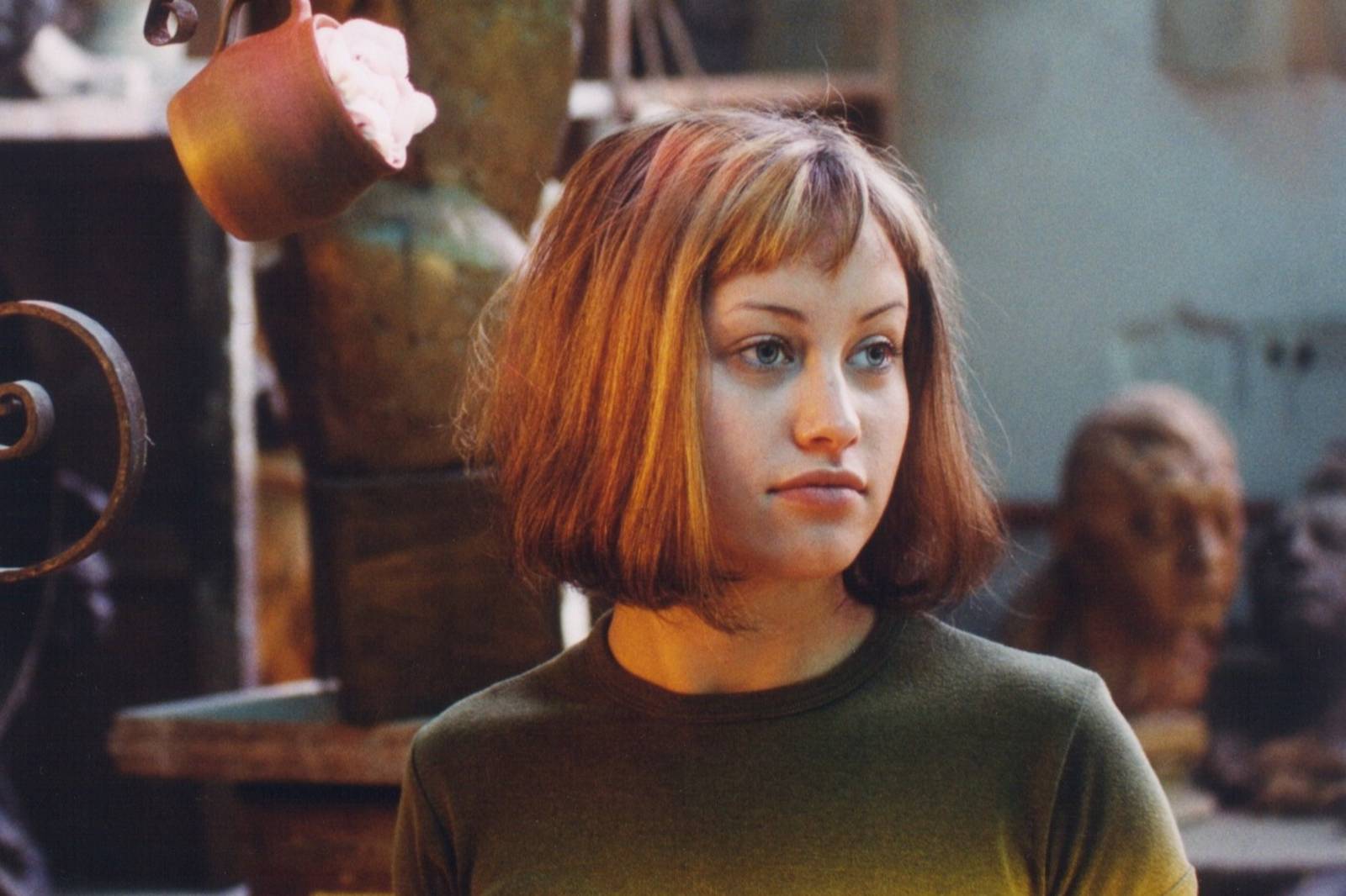 Young woman with a shoulder-length brown bob, olive-green top, looking to the right; blurred sculpture background.