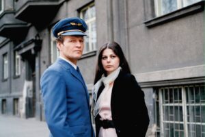 A man in a blue pilot uniform and hat stands beside a woman with dark hair in a pale pink top and black coat on a city sidewalk.