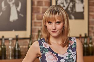 Smiling woman with blonde hair in a floral print dress poses in a bar or cafe setting with bottles in the background.