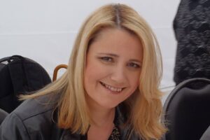 Close-up of a smiling blonde woman wearing a black leather jacket and a leopard-print top, looking at the camera.