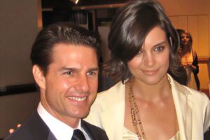 Smiling man in a tuxedo and a woman in a cream blazer posing together at a formal event. The woman wears layered gold necklaces and has shoulder-length brown hair.