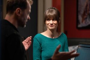 Woman in a teal top smiles while conversing with a man in a dimly lit room; a laptop is visible on the table nearby.