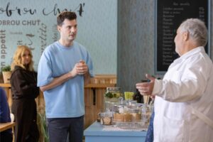 Man in a light blue shirt speaks with an elderly man in a white coat at a cafe counter, with cakes on display behind them.