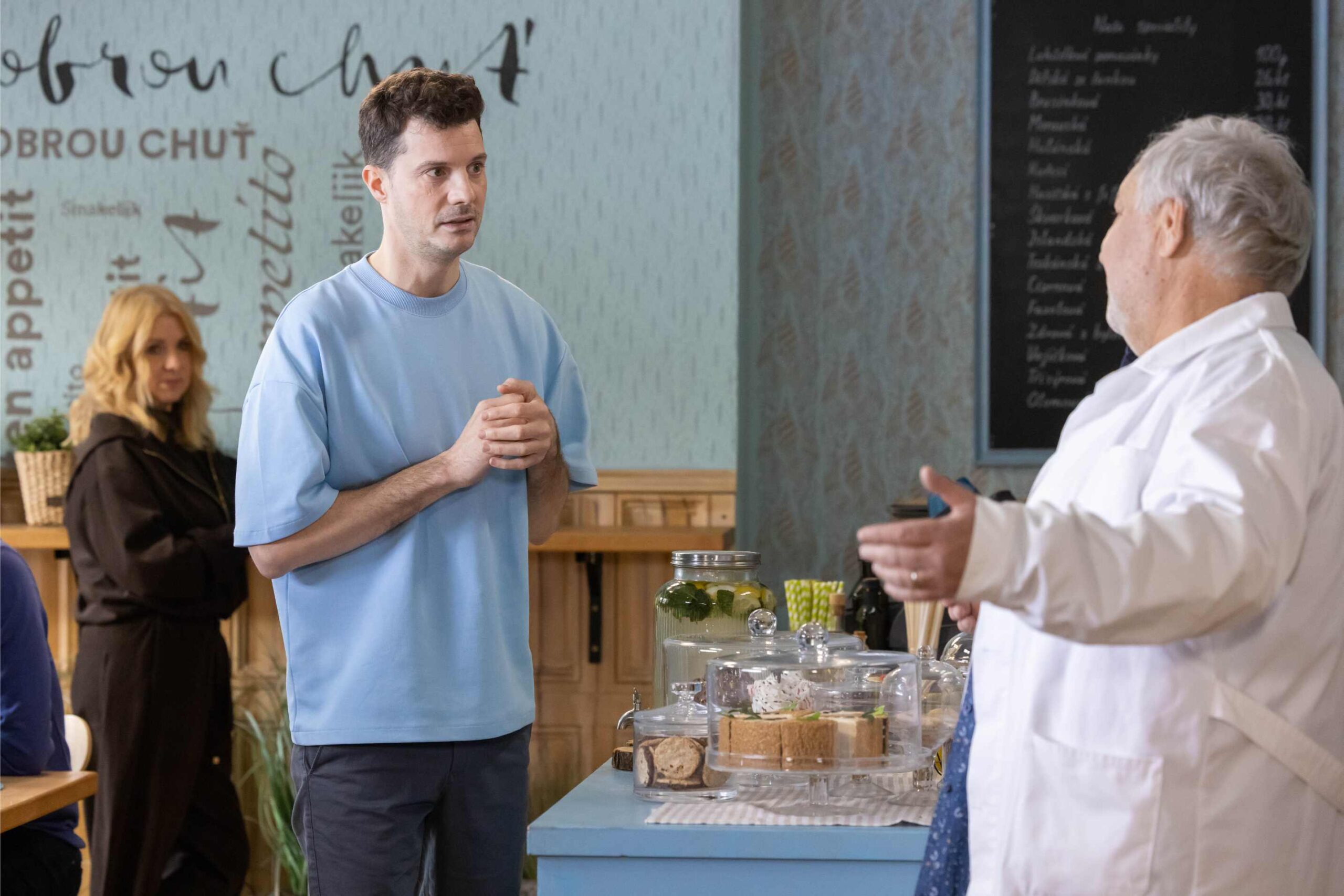 Man in a light blue shirt speaks with an elderly man in a white coat at a cafe counter, with cakes on display behind them.