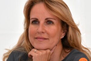 Close-up of a middle-aged woman with blonde hair resting her chin on her hands, looking thoughtful.