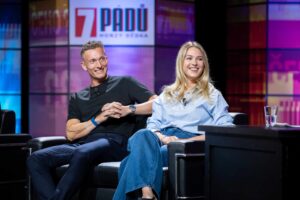 Two presenters seated on a panel stage, smiling and chatting against a colorful set backdrop.