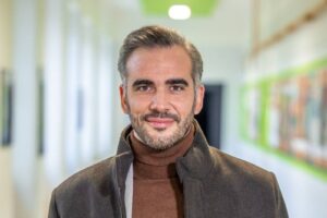 Smiling man with gray hair and stubble wearing a brown turtleneck and dark coat in a bright hallway.