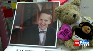 Framed portrait of a smiling man in a tuxedo beside a teddy bear holding a pink heart, part of a memorial display on a news broadcast.
