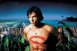 A shirtless man with a red swirl painted on his chest, wearing a green stone necklace, stands among ropes and tropical plants at night under a starry sky.