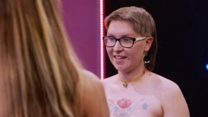 Person with short hair and glasses speaks to someone off-camera, wearing a rainbow heart necklace and floral chest tattoo.