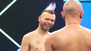 Smiling man with a blond mohawk and a circular chest tattoo, wearing a silver necklace, conversing with a second shirtless man whose back is to the camera.