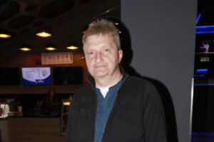 Man with short blond hair and stubble smiles at the camera in a dimly lit cafe or bar setting, wearing a dark jacket over a blue shirt.