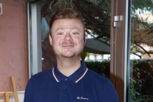 Smiling man with light brown hair and stubble wearing a navy polo, standing indoors beside a glass door.