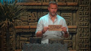 Man in a light blue shirt holds a torn brown sign with the word STANDA in front of a carved stone backdrop.
