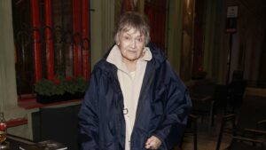 Older woman in a navy raincoat and beige zip sweater stands on a dim city street at night, with a neutral expression.