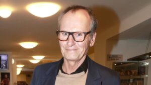 Smiling, middle-aged man with large brown-framed glasses, wearing a tan sweater and dark blazer in a warmly lit hallway or gallery.