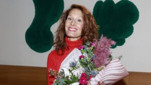 Smiling woman in a red turtleneck holding a bouquet of pink and purple flowers against a wall with green abstract shapes behind her.