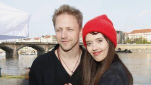 Smiling couple posing near a river with a stone bridge and city buildings in the background, on a sunny day.