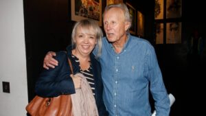 Smiling woman and older man pose together in an art gallery, arms around each other as framed artwork hangs in the background.