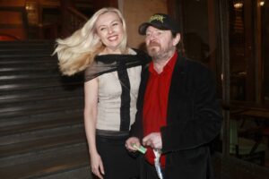 Smiling blonde woman in a beige dress with a black scarf posing with a bearded man in a red shirt and cap outside a building.