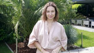 Woman in a light pink blouse posing outdoors among lush green palm plants, smiling softly.