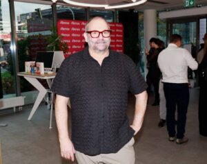 Man with red-framed glasses and a patterned black shirt posing at an event with an Alensa backdrop in the background.