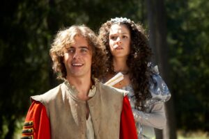 Two people in period costumes outdoors; man in red and beige tunic smiles in the foreground, woman in a silver gown stands behind him.